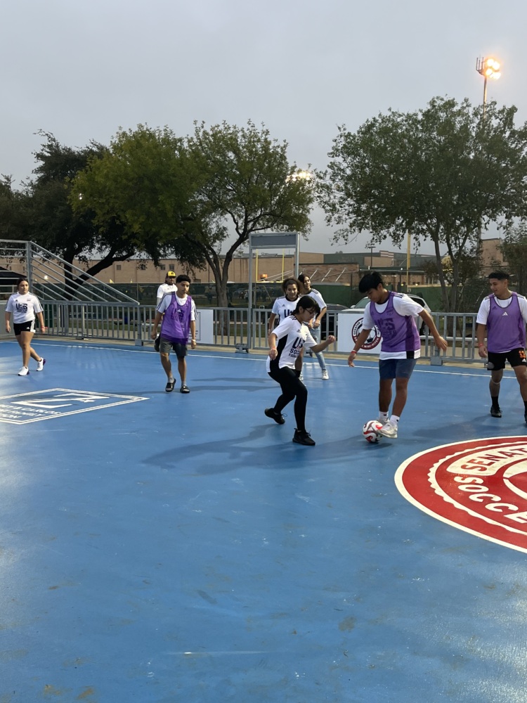 A group of kids half in white shirts and half in purple pinnies play soccer on a blue mini-pitch